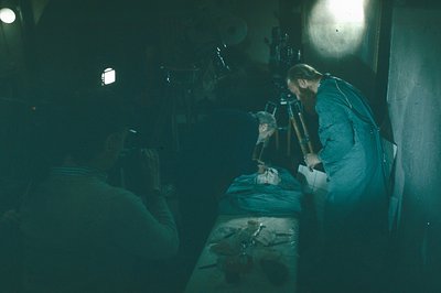 A man with a beard leans over a prone figure covered by a sheet, appearing to perform a medical procedure in a dimly lit room...