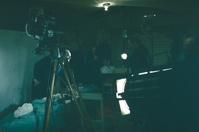 A film set scene: a vintage 16mm camera mounted on a tripod dominates the foreground. Figures are partially visible in shadow...
