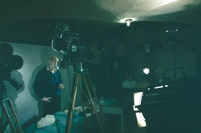 A film set interior, likely a studio or basement, features a cameraman adjusting a vintage film camera on a tripod. Visible c...