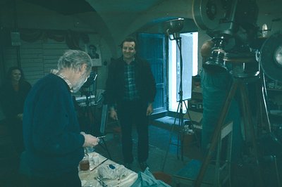 A man with a focused expression meticulously works on a sculpture in a cluttered studio. A second man in a plaid shirt stands...
