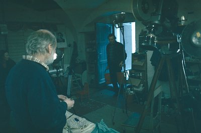 An older man, seated and working on a sculpture, is the focus in a cluttered, dimly-lit workshop. A large-format camera and l...