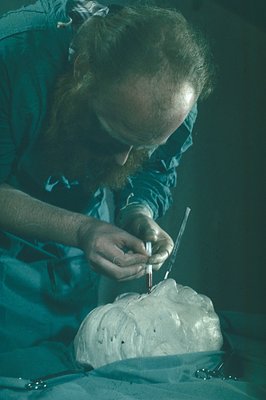 A sculptor with a full beard, wearing a lab coat, meticulously applies pigment to a plaster bust, likely during restoration. ...