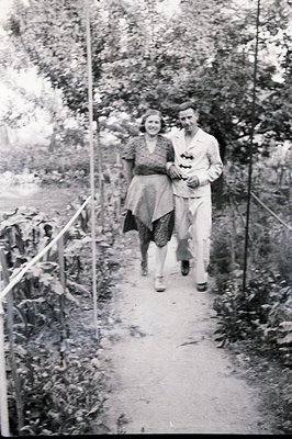 A couple strolls along a narrow, gravel path lined with vegetation. The woman wears a V-neck dress with a prominent sash; the...