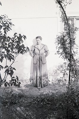 Formal portrait of a woman in a patterned, high-waisted dress, posed outdoors against a white wall & overgrown trellis. Appea...