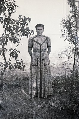 Full-length portrait of a woman in a long-sleeved, patterned dress, standing outdoors. Lush greenery and a stone wall form th...