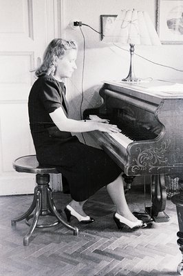 A woman in a dark dress and pumps sits at an ornate, upright piano. Interior view shows a well-appointed room with hardwood f...
