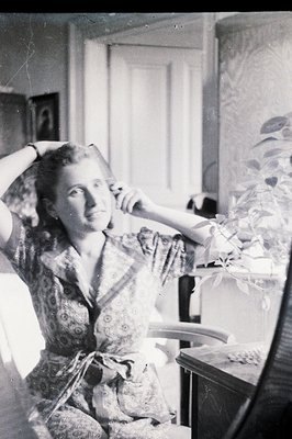 A seated woman in a patterned dress poses with her hands raised, appearing to adjust her hair. Interior scene with a patterne...