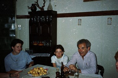 Three people sit at a table laden with pastries, likely a celebratory meal. The interior features a dark wood sideboard and p...