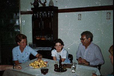 A family gathers around a table laden with food, likely enjoying a meal. Three individuals are visible – a woman in a blue sh...