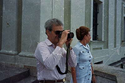 Man with a camera captures a moment alongside a woman in a 1970s-style blue dress. Architectural facade with classical detail...