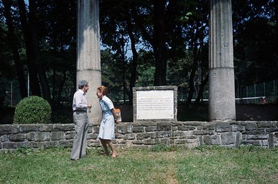 A man and woman, likely tourists, engage in conversation near a monument featuring two classical columns and a stone plaque. ...
