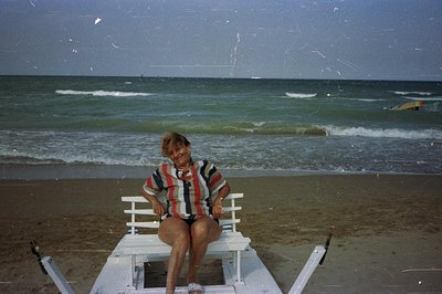 A woman sits on a white, folding beach chair facing the camera. She wears a patterned, short-sleeved top and shorts. Behind h...