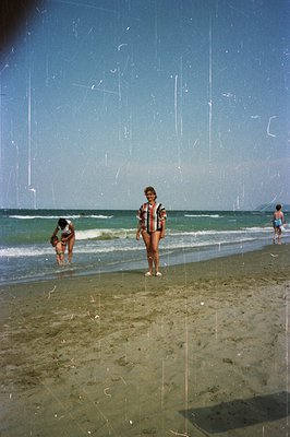 Seaside snapshot: A woman in a checked swimsuit stands on a sandy beach, facing the camera. Two children play in the surf nea...