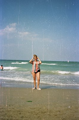 A young woman in a vintage, patterned swimsuit and short sleeves stands on a sandy beach, appearing to pose for the camera. G...