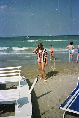 A woman in a patterned swimsuit stands on a sandy beach, casting a shadow near white lounge chairs. Visible in background are...