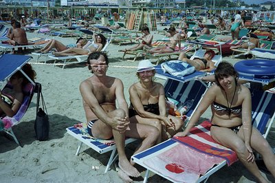 Three people seated on beach chairs at a crowded sandy shoreline. Visible details include patterned towels, dark swimsuits, a...