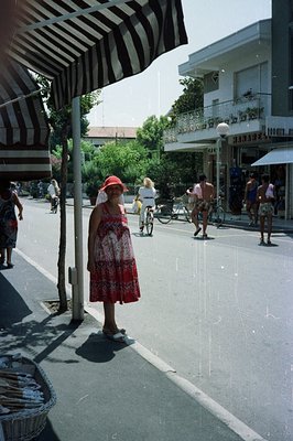 A woman in a floral dress & straw hat stands near a striped awning on a sun-drenched street lined with shops and bicycles. Me...