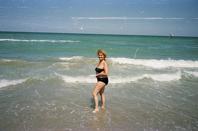A woman stands knee-deep in the shallow waters of a beach, appearing to enjoy the waves. She wears a black bikini and has sho...