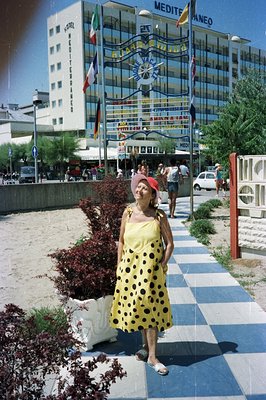 A woman in a yellow, polka dot dress and hat poses on a patterned walkway beside a beachfront hotel. The hotel boasts a mid-c...