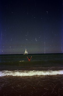 A person in an orange swimsuit extends arms as they enter waves near a sailboat under a dark sky. Grainy image suggests film ...