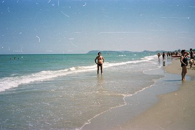 A man wades in shallow, turquoise water along a sandy beach. A woman stands nearby, both figures appearing casual. Crowd visi...