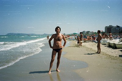 A man stands at the water’s edge on a crowded beach. He wears black swim trunks and has short, dark hair. Numerous beachgoers...