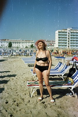 A woman stands on a sandy beach wearing a black bikini and a pink sunhat. Rows of beach chairs extend into the background, wi...
