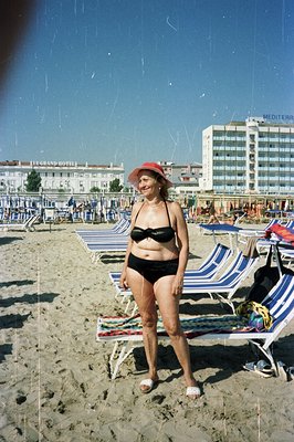 A woman in a black bikini and wide-brimmed hat poses on a sandy beach with rows of striped deckchairs extending into the back...