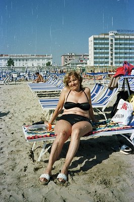 A woman in a black bikini sits on a beach chair, surrounded by other patrons and rows of striped umbrellas. A high-rise hotel...
