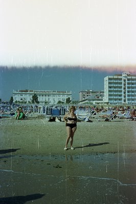 Seaside resort scene: A woman in a black bikini stands on a sandy beach with sunbathers and a hotel in the background. Likely...