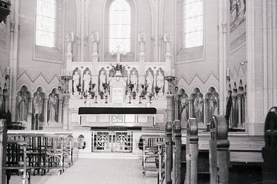 Ornate, black and white photo of a Catholic church interior. Focus on the altar, adorned with floral arrangements & statues. ...