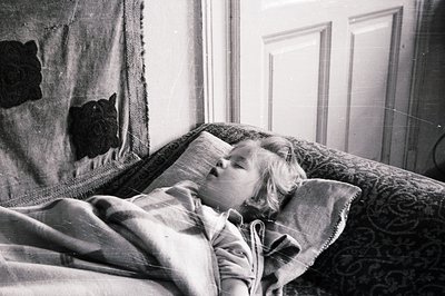 A young child sleeps peacefully, bundled in a blanket, within a bedroom. Detailed wallpaper and a dark floral throw provide r...