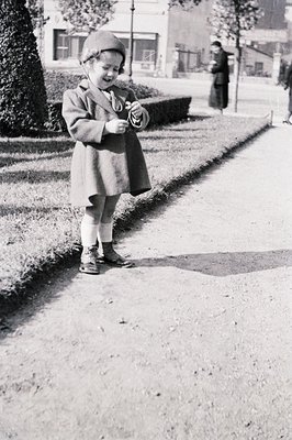 A young child, bundled in a wool coat and hat, stands near a manicured hedge. White stockings and sturdy shoes complete the o...