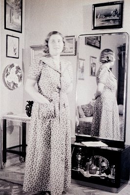 Full-length portrait of a woman in a patterned dress standing before a tall, ornate mirror. Interior setting with decorative ...
