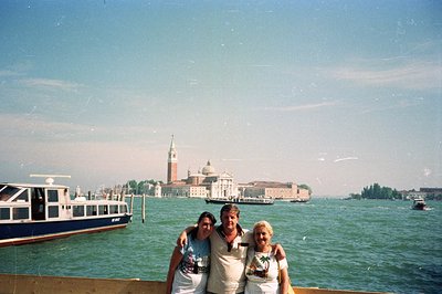 A family of three poses for a photo with San Giorgio Maggiore island and Venice in the background. The subjects, likely touri...
