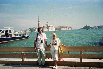 A couple stands near a weathered railing, holding hands, with San Giorgio Maggiore island visible across the Venetian lagoon....