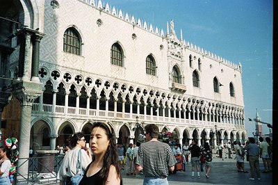 An exterior view of the Doge's Palace in Venice, Italy, featuring its iconic arcaded facade. A young woman stands in the fore...