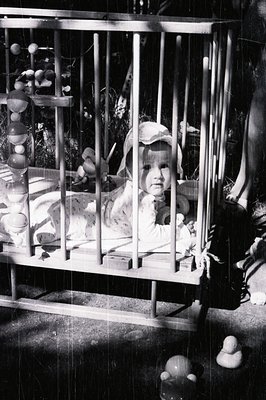 Infant lies within a vintage wooden crib, gazing directly at the camera. A mobile with wooden beads hangs overhead. Likely ta...