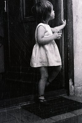 A young child in a short dress and sandals stands on a welcome mat, gently touching a door frame. The image, likely from the ...