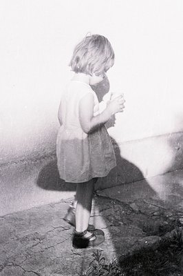 A young girl in a simple dress and Mary Jane shoes examines something held in her hands against a textured wall. The photo's ...