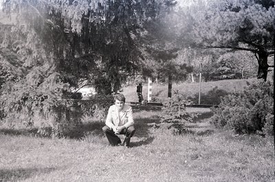 A young man, dressed in a light-colored collared shirt and dark trousers, squats within a grassy area, flanked by dense folia...