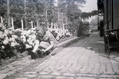 A woman in a 1940s-style dress kneels, tending a flower bed along a brick-paved walkway. A weathered wooden fence and overgro...
