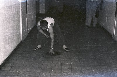 A man in a sleeveless shirt and sandals appears to be gesturing emphatically on a tiled floor within a narrow hallway or corr...