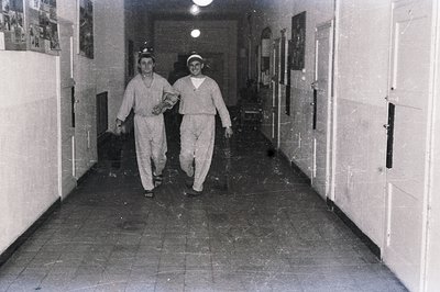 Two men in uniform and headgear walk down a tiled hallway. The scene appears to be an institutional setting, possibly a hospi...