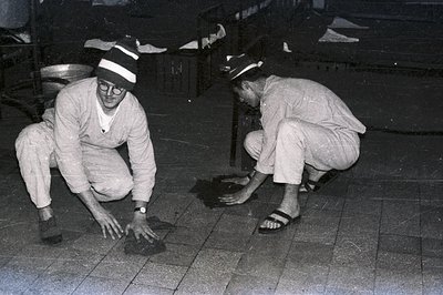 Two men in what appears to be hospital attire and caps kneel, examining a dark object on a tiled surface. Likely a medical or...