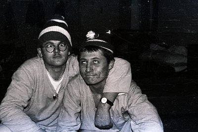 Two men wearing knit hats sit close together; one with glasses and a white t-shirt, the other with a visible wristwatch. The ...