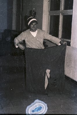 A man in a dark sweater and spectacles poses with a large, dark fabric draped over his body, seemingly held up by his arms. V...