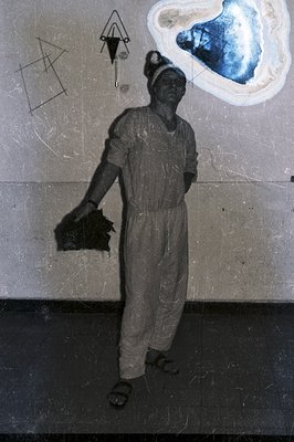 A man in a jumpsuit stands in a stark, indoor space, seemingly a workshop or studio. He holds a dark object and wears protect...