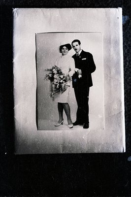 Black and white portrait of a formally dressed couple, likely a wedding photo. The woman wears a short dress and hat, carryin...