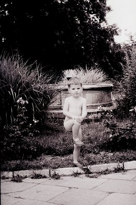 A young boy, barefoot, sits with legs crossed, examining a scraped knee. Dense greenery and a stone wall form the backdrop. T...